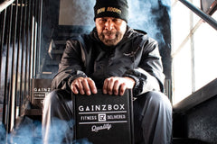 This image shows a middle aged BIPOC man sitting on the stairs surrounded by thematic smoke holding a closed Gainz Box.