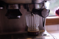 Espresso machine pouring shots of espresso into a clear glass swirly mug, a window in the background