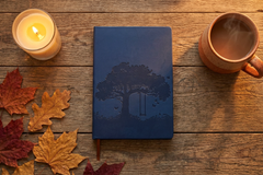 Navy 'Tree of Life' hardcover journal with an embossed tree design, flat-lay on a wood table with a candle, coffee, and autumn leaves.