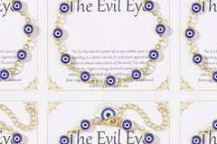 Detail close-up of a single 14K gold plated evil eye bracelet on 'The Evil Eye' gift card, showing blue evil eye charms on a gold-tone chain