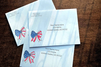 Three white envelopes with printed U.S. mailing addresses and small red, white and blue patriotic bow designs on a wooden table.