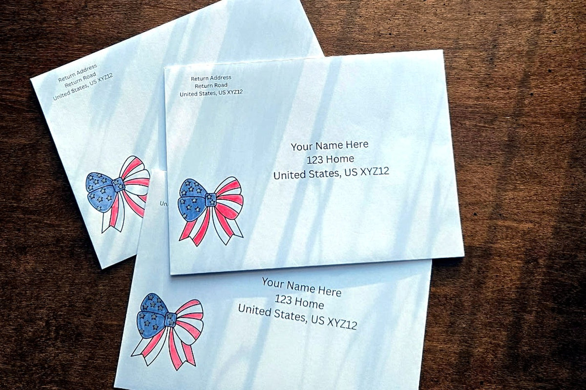 Three white envelopes with printed U.S. mailing addresses and small red, white and blue patriotic bow designs on a wooden table.
