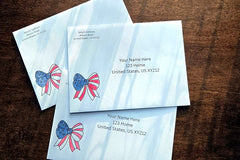 Three white envelopes with printed U.S. mailing addresses and small red, white and blue patriotic bow designs on a wooden table.