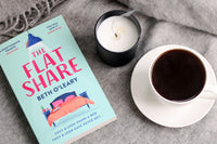 A book called the flat share laying on a piece of cloth or a blanket with a coffe mug and a white candle next to it.