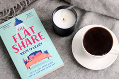 A book called the flat share laying on a piece of cloth or a blanket with a coffe mug and a white candle next to it.