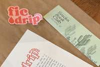 Several items laid out on top of a brown paper shipment bag, including a pink and red FicDrip vinyl sticker, and a bookmark.