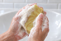 Hands working the pale olive-yellow Turmeric & Black Pepper tallow bar into a thick white lather over a white washbasin