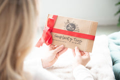Image of woman holding first package of Simplicity Teas of the month delivery. Red Ribbon around package for the holidays. Looks cozy.