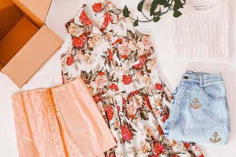 cottagecore flat lay outfit with floral dress, pink corset, high waisted denim and folded white top with a box