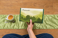 Lifestyle shot holding the Tea Smith tea of the month box, sitting on a green satin pillow on a wood floor with a cup of tea. 