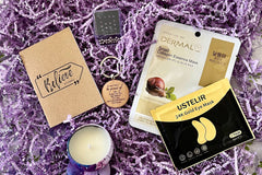 Close up look to Purple Box self-care items, such as scented candle, keychain, notepad, fidget toy, eye mask and face mask.