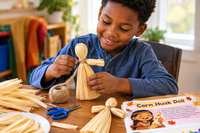 A smiling young boy in a blue shirt makes a corn husk doll at a wooden table, carefully arranging the dried husks and tying them with twine