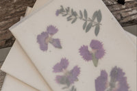 A set of 3 cards in clear paper envelopes laying on an old wooden picnic table. Each card is decorated with pressed wildflowers.