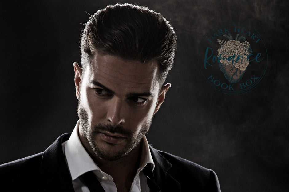 Handsome man standing with his chin pointed down and looking away. Dark Heart Romance logo to the right of him. Dark background