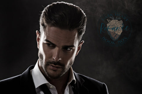 Handsome man standing with his chin pointed down and looking away. Dark Heart Romance logo to the right of him. Dark background
