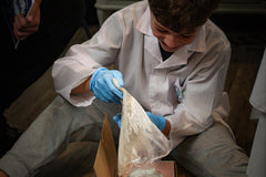 A child in a lab coat and gloves is on the floor, pouring plaster into a foot impression made in biofoam. 