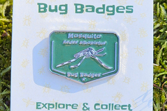Close-up of a Mosquito enamel pin (green) on a Bug Badges backing card, shot outdoors on moss with the “Bug Badges”
