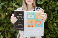 A woman holds up 'The Pivot Year' and a You Should Read mailer outdoors with lush green foliage in the background.