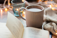 AN OPEN BOOK LIES ON A TABLE. A CUP OF HOT COFFEE SITS CLOSE BY. WHITE CHRISTMAS LIGHTS ARE STRUNG ALONG IN THE BACKGROUND.