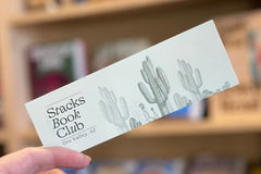 Close-up of a seafoam green bookmark with a desert scene sketched onto it and the Stacks Book Club logo.