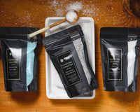 Introverts Read & Relax box contents. Three bags of bath salts on a wooden table. One bag is opened an on a tray.