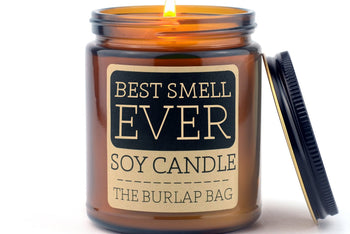 Lit Best Smell Ever 9oz soy candle by The Burlap Bag in an amber glass jar, black metal lid resting beside it on white