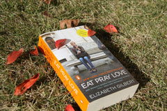 Close up photo of a romance book called eat the pray love laying on grass outside and with leaves on it and next to it.