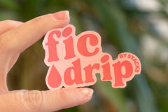 Close-up of two fingers holding a pink and red vinyl sticker that says FicDrip by Stacks with a droplet icon.