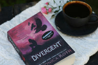 Close up photo of a famous book called divergent from Roth and a cup of liquid while all being on a white napkin