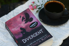 Close up photo of a famous book called divergent from Roth and a cup of liquid while all being on a white napkin