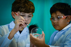 Two children in lab coats and safety glasses are dusting a glass for fingerprints. One is holding the glass and the other is dusting. 