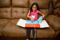 A delighted girl  opens a StoryCaptain Books Club box, her eyes wide with excitement as she discovers a collection of new picture books