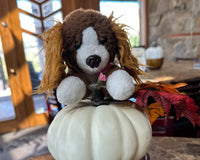 A brown and white dog sitting ontop of a white pumpkin. The dog is a stuffed animal. You can see the lighter brown ears.