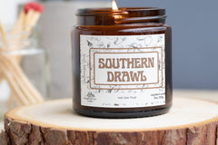 Southern Drawl candle. Amber jarred candle sitting on a slice of wood. Red-tipped matches stand in a jar in the background.