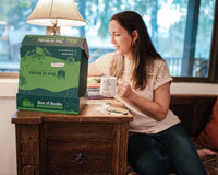Woman at home, peeking into a Books & More by Box of Books while enjoying a cuppa. Box of Books specializes in great reads and novelty items