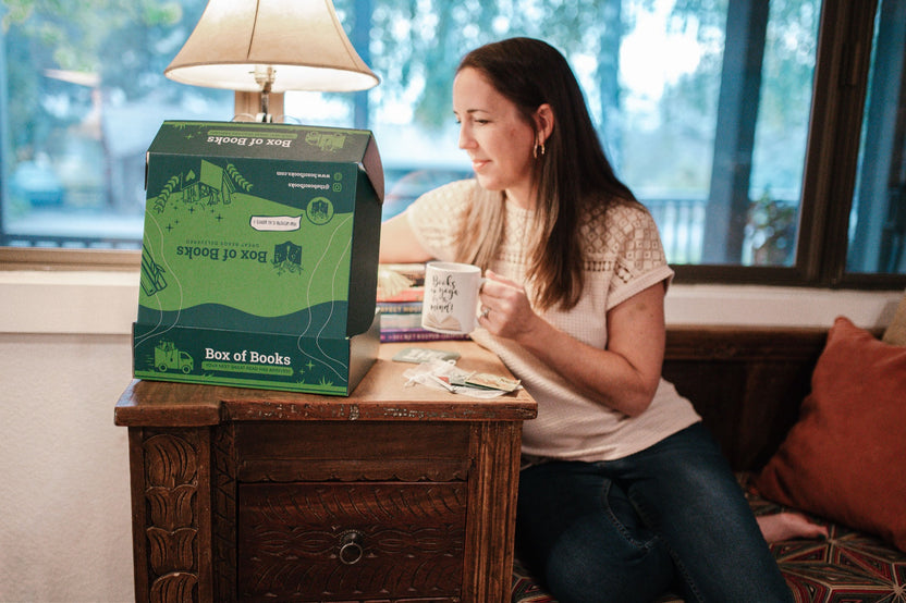 Woman at home, peeking into a Books & More by Box of Books while enjoying a cuppa. Box of Books specializes in great reads and novelty items