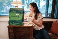 Woman enjoying a cup of tea while looking at a Box of Books subscription box on a table by the window