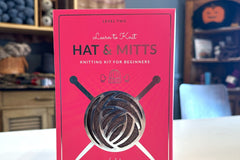Learn to Knit Hat & Mitts knitting kit for beginners - box on tabletop with yarn shop background - brown yarn showing through the box