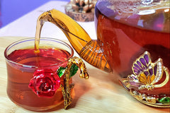 A zoomed in shot of a teapot pouring my steeped Walk in the Woods tea into a small, glass tea cup on the left.