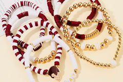 Two piles of loose Game Day stackable friendship bracelets on a cream surface, one in maroon and white beads and one in gold beads, with foo