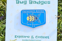 Close-up of a Cucumber Beetle enamel pin on a Bug Badges backing card, photographed on bright green moss outdoors