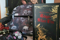 Black skull and red flower backpack in front of a rose picture, with a raven and skull statue, Edgar Allan Poe book, and glasses case