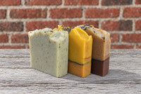 Three of SoapyFluffs' hand-cut soap bars, two of them layered, topped with seeds and botanicals, on a weathered wood table