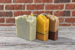 Three of SoapyFluffs' hand-cut soap bars, two of them layered, topped with seeds and botanicals, on a weathered wood table