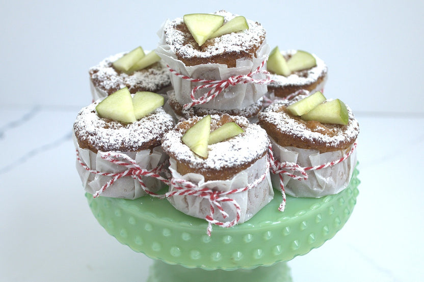 Mini golden apple cakes studded with apple bits, dusted with sugar, alongside muffin pan and baking tools