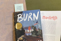 A hardcover copy of Burn by Peter Heller with a "signed first edition" sticker on it, with a bookmark poking out of the top, beside a flyer,