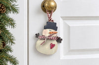 The wooden snowman ornament hanging from its gingham ribbon loop on the brass knob of a white farmhouse door beside fir garland