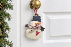 The wooden snowman ornament hanging from its gingham ribbon loop on the brass knob of a white farmhouse door beside fir garland