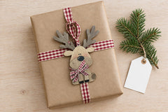 The wooden reindeer ornament tied onto a kraft-wrapped gift with red gingham ribbon as a gift topper, beside a fir sprig and tag