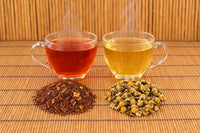 Cinnamon Sensation Rooibos brewed red-amber and Chamomile with Lemon brewed pale gold in glass cups, each beside its loose tea